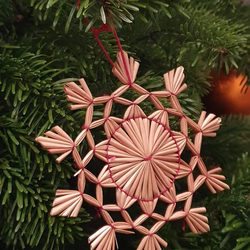 Eine kunstvoll gestaltete Schneeflocke aus Stroh hängt an einem grünen Tannenbaum. Der warme Glanz des Materials vermittelt eine festliche Atmosphäre.