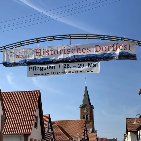 Ein historisches Dorffest mit einem großen Banner über der Straße. Im Hintergrund ist eine Kirche und hübsche, traditionelle Gebäude zu sehen.