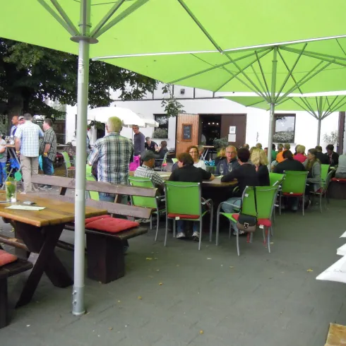 Ein gemütlicher Außenbereich mit vielen Gästen unter grünen Sonnenschirmen. Tische sind mit bunten Stühlen umgeben, was eine einladende Atmosphäre schafft.