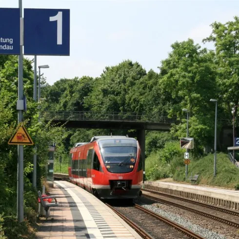 Ein roter Zug fährt an einem Bahnhof mit dem Schild "Richtung Landau" vorbei. Die Umgebung ist grün und von Bäumen umgeben.