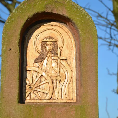 Eine kunstvolle Holzschnitzerei zeigt eine Figur mit einem Schwert und einem Rad. Im Hintergrund sind Äste und ein klarer blauer Himmel zu sehen.