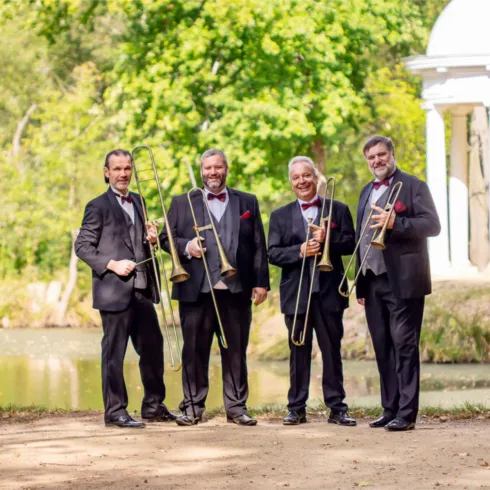 Vier Musiker stehen am Ufer eines Teichs und halten Posaunen in der Hand. Sie tragen elegante Anzüge mit Fliegen und lächeln in die Kamera.
