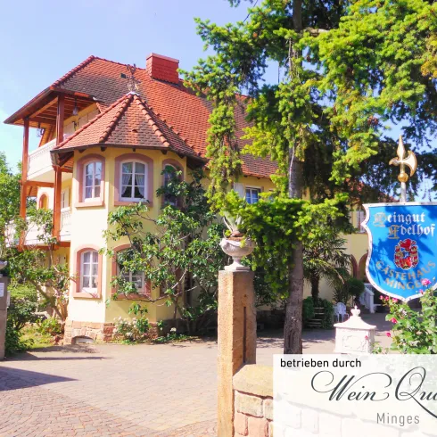 Ein charmantes Gebäude mit einem roten Dach, umgeben von Bäumen und blühenden Pflanzen. Neben dem Eingang steht ein blaues Schild mit einem Wappen.