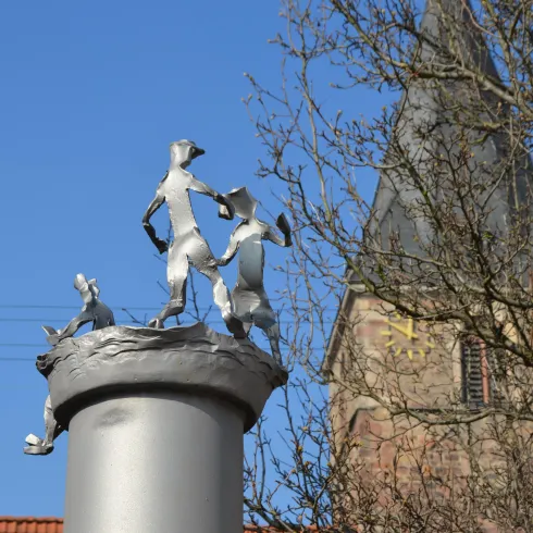 Eine moderne Skulptur von tanzenden Figuren auf einem Säule. Im Hintergrund ist ein Kirchturm sichtbar, und der Himmel ist klar und blau.