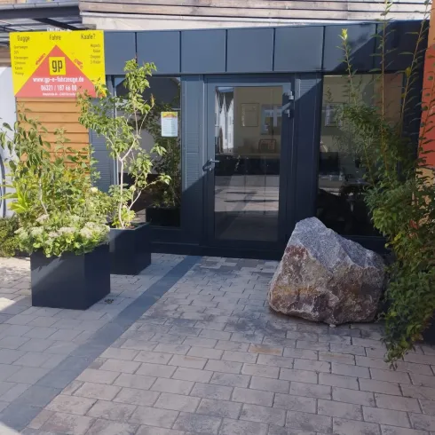 Ein moderner Eingangsbereich mit großen Fenstern und Pflanzen in schwarzen Kübeln. Der Boden ist aus Pflastersteinen, und ein großer Stein liegt dekorativ auf dem Weg.