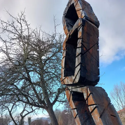 Eine abstrakte Holzskulptur steht in einer Wiese, umgeben von einem Baum und grünem Gras. Der Himmel ist teilweise bewölkt und verleiht der Szene eine ruhige Atmosphäre.