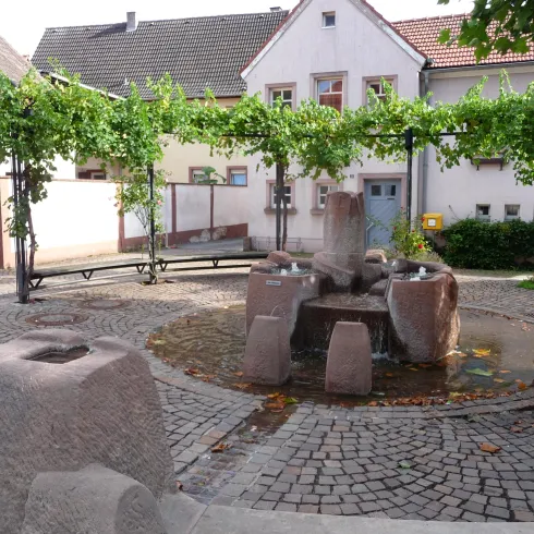 Ein schöner Platz mit einem Brunnen und Weinreben. Im Hintergrund sind Häuser zu sehen, die eine gemütliche Atmosphäre schaffen.