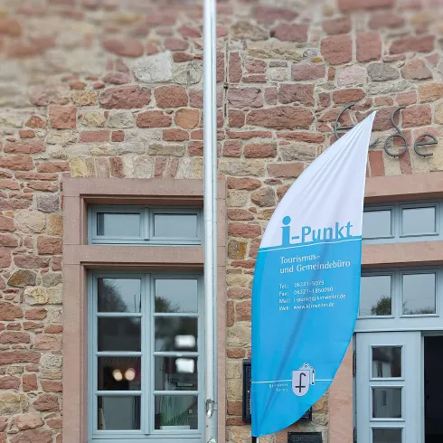 Ein Eingangsbereich eines Gebäudes mit einer Fahne, die den Schriftzug "i Punkt" trägt. Neben der Fahne befinden sich Blumen in einem Pflanzgefäß.