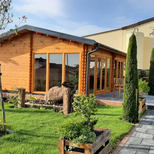 Ein gemütliches Holzhaus mit großen Fenstern steht in einem gepflegten Garten. Umgeben von Bäumen und straff geschnittenen Hecken wirkt die Umgebung einladend und ruhig.