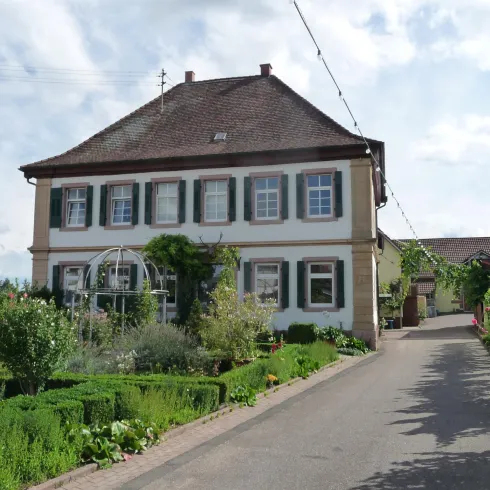 Ein charmantes zweigeschossiges Haus mit grünem Garten und blauen Fensterläden. Der Weg führt zur Eingangstür und ist von einer wunderschönen Landschaft umgeben.