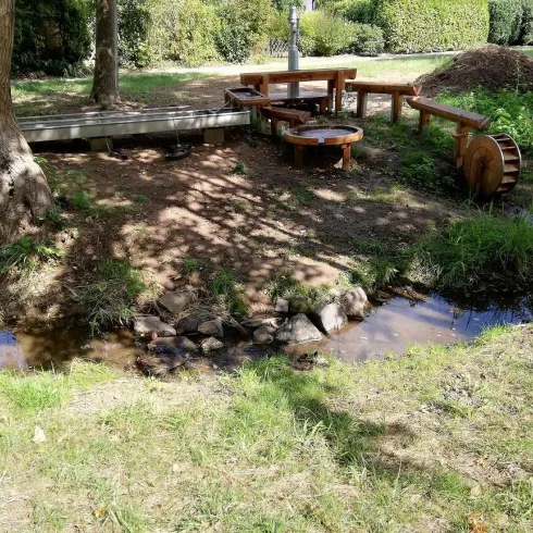 Ein ruhiger Gartenbereich mit einem kleinen Bach und Holzstühlen. Im Hintergrund sind Bäume und ein Haufen Erde zu sehen.