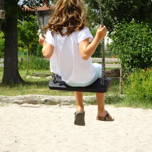 Ein Mädchen schwingt auf einer Schaukel im Sandspielbereich. Die Umgebung ist grün und sonnig.