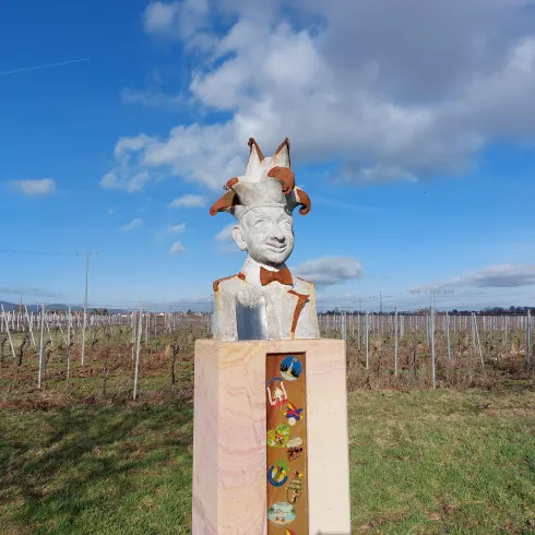 Eine Statue mit einem humorvollen Gesicht und einem lustigen Hut steht in einem Weinberg. Im Hintergrund sind Reben und ein klarer blauer Himmel zu sehen.