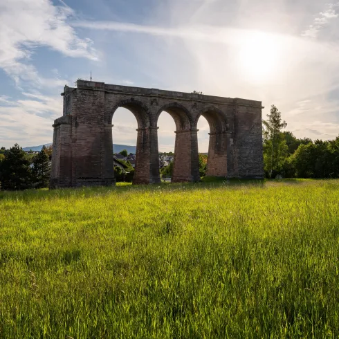 Eine alte Ruine mit Bögen steht in einem grünen Feld. Der Himmel ist blau mit einigen Wolken und die Sonne scheint.