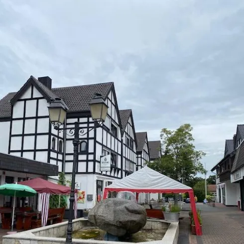 Ein malerischer Platz mit Fachwerkhäusern und einem Brunnen. Markisen und Tische laden zum Verweilen ein.