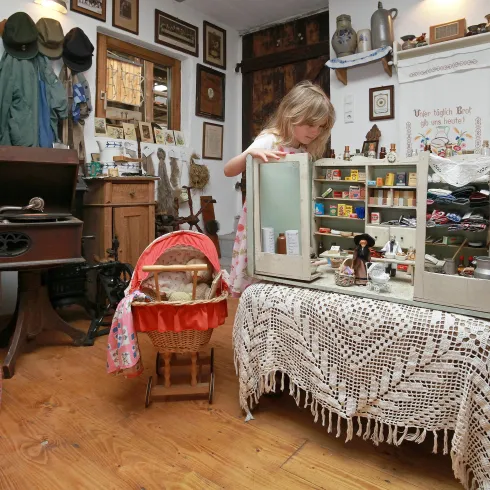 Ein nostalgischer Raum mit einem Kind, das an einem Puppenhaus spielt. Im Hintergrund sind alte Möbel und verschiedene Ausstellungsstücke zu sehen.