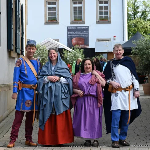 Vier Personen in historischen Kostümen stehen auf einem gepflasterten Weg. Im Hintergrund sind kleine Stände und eine alte Fassade zu sehen.