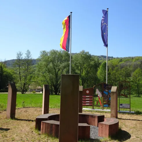 Ein Platz mit Flaggen von Deutschland, der EU und Frankreich. Im Hintergrund sind Bäume und eine Wiese zu sehen.