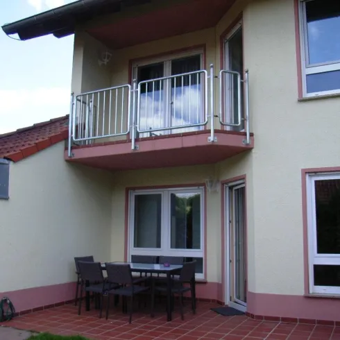 Ein modernes Haus mit einem Balkon und einem kleinen Essbereich im Freien. Die Fassade ist hell bemalt und der Himmel ist teilweise bewölkt.