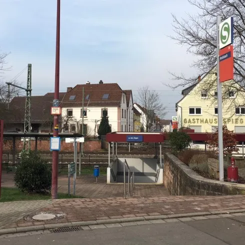 Eine Haltestelle mit einem Informationsschild und dem Gebäude eines Gasthauses im Hintergrund. Die Umgebung ist ruhig mit einigen Bäumen und einem klaren Himmel.