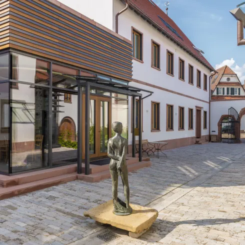 Ein moderner Gebäudeteil mit großen Fenstern und Holzverkleidung. In der Mitte steht eine Statue auf einem Holzsockel, umgeben von einer gepflasterten Fläche und Pflanzen.