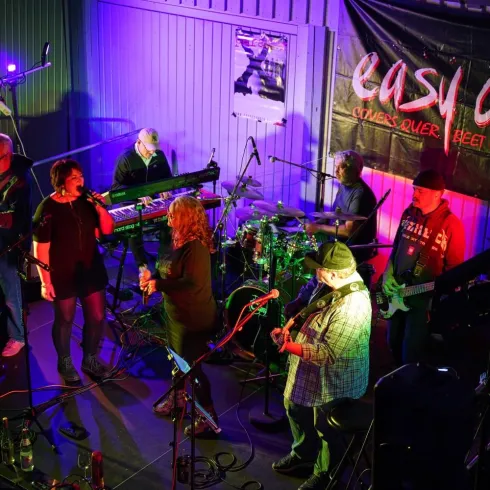 Eine Liveband spielt auf einer Bühne mit bunten Lichtern. Die Musiker scheinen viel Spaß zu haben und das Publikum zu begeistern.