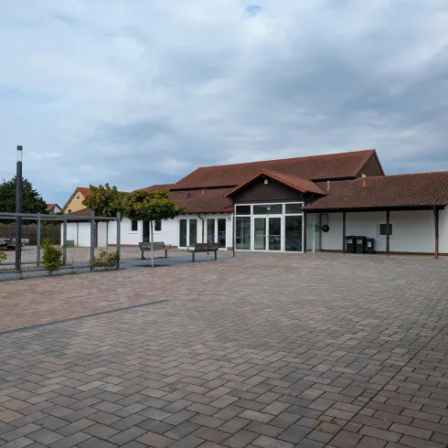 Ein modernes Gebäude mit einem großzügigen Platz davor. Der Himmel ist bewölkt und es gibt einige Bäume und Sitzbänke in der Umgebung.