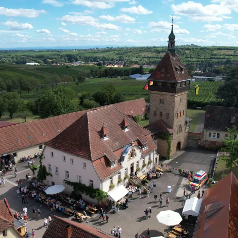 Ein historisches Gebäude mit einem hohen Turm und roten Dächern. Der Innenhof ist belebt und von einer schönen Landschaft umgeben.