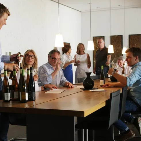 Eine Gruppe von Menschen sitzt an einem langen Tisch und genießt Wein. Einige Personen sprechen miteinander, während andere den Wein betrachten.