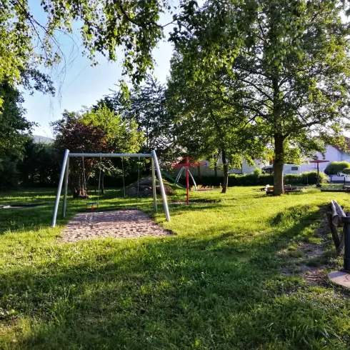Ein Spielplatz mit einer Schaukel und einer Rutsche, umgeben von hohen Bäumen. Die Wiese ist grün und einladend.