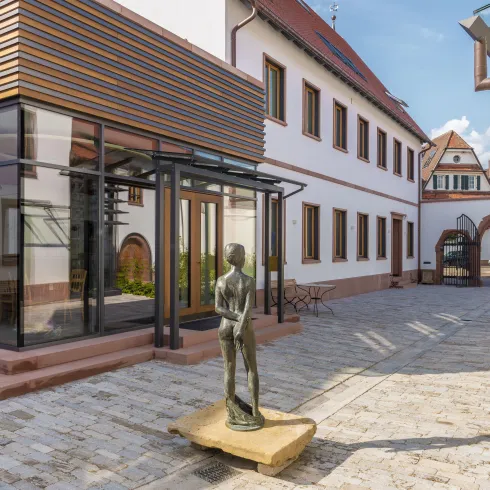 Ein moderner Innenhof mit einem Glasgebäude und einer Skulptur. Im Hintergrund sind traditionelle Gebäude im regionalen Stil zu sehen.