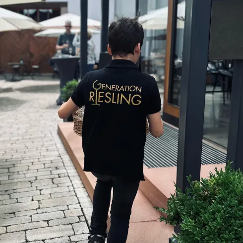 Ein Junge trägt ein schwarzes T-Shirt mit dem Aufdruck „Generation Riesling“ und hält einen Korb. Im Hintergrund sind Tische und Sonnenschirme zu sehen.