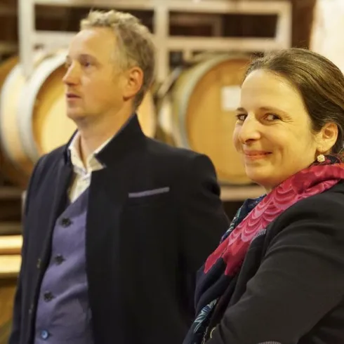 Ein Mann und eine Frau stehen in einem Weinkeller zwischen Eichenfässern. Die Frau lächelt freundlich, während der Mann nachdenklich aussieht.