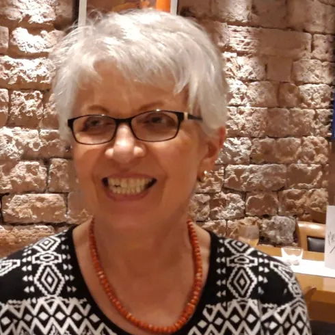 Eine lächelnde Frau mit kurzen, grauen Haaren und Brille sitzt in einem gemütlichen Restaurant. Der Hintergrund besteht aus Ziegelmauern.