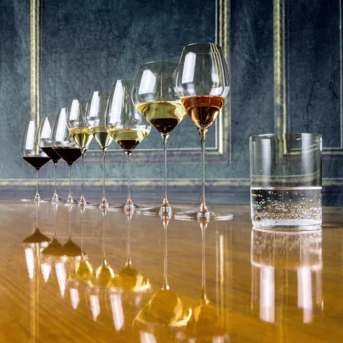 Eine Reihe von Riedel Weingläsern mit verschiedenen Weinen stehen auf einem Holztisch. Neben den Gläsern befindet sich ein Glas mit Wasser.