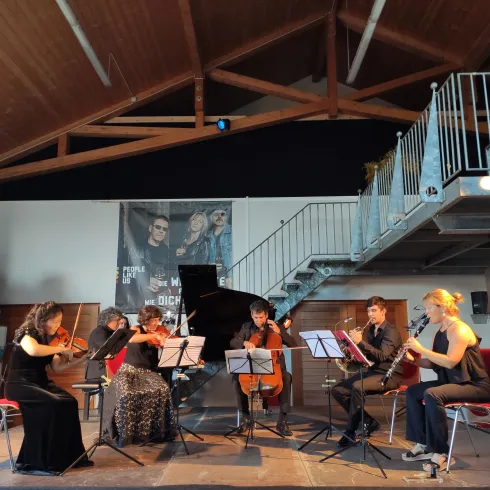 Ein Ensemble aus sechs Musikern spielt in einem modernen Raum. Im Hintergrund sind große Poster und ein Klavier zu sehen.