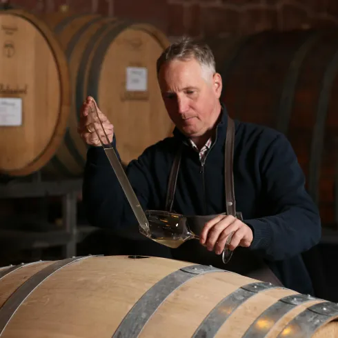 Oenologe Thorsten Ochocki nimmt mit dem gläsernen Stechhaber Weißwein aus einem Holzfass, dem Barrique. Im Hintergrund sind weitere Fässer sichtbar, Weinkeller-Atmosphäre.