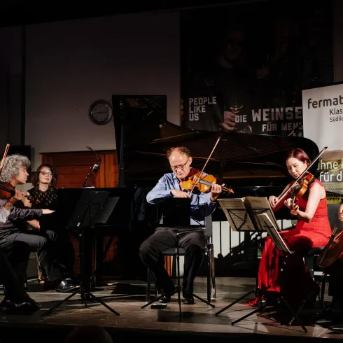 Ein Ensemble aus fünf Musikern spielt klassische Musik auf der Bühne. Im Hintergrund sind ein Klavier und ein Banner zu sehen.