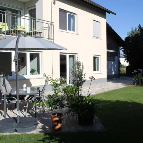 Ein hübscher Garten mit einer Terrasse, einem Tisch und Stühlen. Im Hintergrund sieht man ein modernes Haus und einen blauen Himmel.