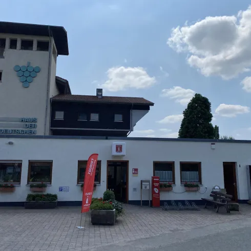 Ein modernes Gebäude mit einer weißen Fassade und einem hohen Turm steht im Vordergrund. Der Himmel ist klar mit einigen Wolken und es gibt eine rote Fahne am Eingang.