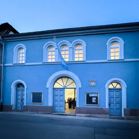 Ein blaues Gebäude mit großen Fenstern und zwei Eingangstüren. Die Fassade ist einfach und modern, mit einem leichtem Lichtschein im Inneren.