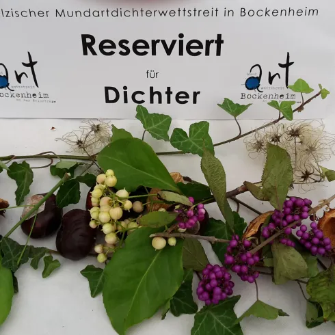 Ein Tisch mit einer Reservierung für Dichter und einer Vielzahl von Kräutern und Blumen dekoriert. Im Hintergrund steht ein Schild mit dem Hinweis auf einen Mundartwettbewerb in Bockenheim.