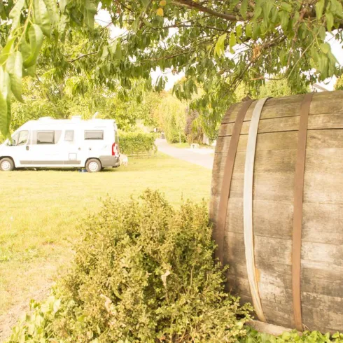 Ein Wohnmobil steht auf einer grünen Wiese neben einem großen Holzfass. Im Hintergrund sind Bäume und eine ruhige Straße zu sehen.