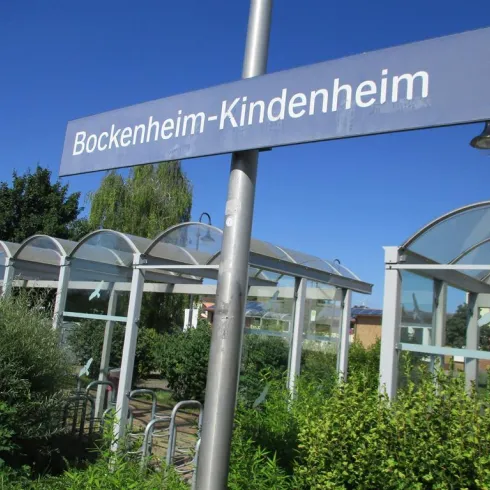 Ein Bahnhofsschild mit der Aufschrift "Bockenheim-Kindenheim". Im Hintergrund sind Bäume und eine moderne Unterstandstruktur zu sehen.