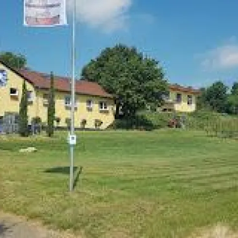 Ein gelbes Gebäude mit einem großen Garten und Weinreben im Hintergrund. Der Himmel ist klar und blau.
