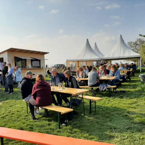 Eine belebte Freiluftveranstaltung mit vielen Menschen an Tischen im Grünen. Im Hintergrund befinden sich ein Verkaufsstand und Zelte.