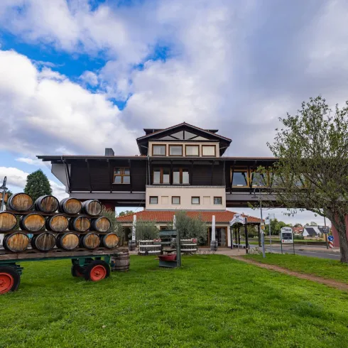 Ein traditionelles Gebäude mit mehreren Etagen und einem großen Balkon. Im Vordergrund stehen Holzfässer auf einem Wagen und viel grünes Gras.