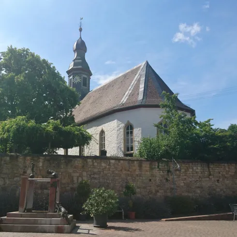 Eine kleine Kirche mit einem charakteristischen Turm, umgeben von Bäumen und einem Mauerwerk. Der Himmel ist blau mit einigen Wolken.