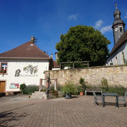 Ein malerischer Platz mit einem historischen Gebäude und einer Kirche. Der Himmel ist klar und blau, umgeben von üppigem Grün.