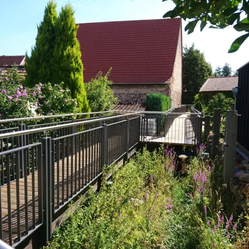 Ein schmaler Holzsteg führt durch grüne Vegetation zu einem Haus mit rotem Dach. Die Umgebung ist hell und sonnig, mit wenigen Gebäuden im Hintergrund.